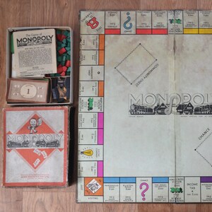 Könnte beinhalten: Ein klassisches Monopoly-Brettspielset bestehend aus einem quadratischen, faltbaren Spielbrett mit traditionellen Immobilienfeldern, einem Kartondeckel mit rotem Rautenmotiv sowie einer Aufbewahrungsbox mit grünen Kunststoffhäusern.
