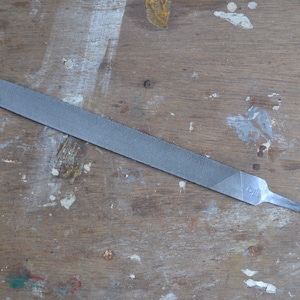 Puede incluir: Una lima de metal plateada con una superficie de dientes finos. La lima está sobre una superficie de madera con salpicaduras de pintura.