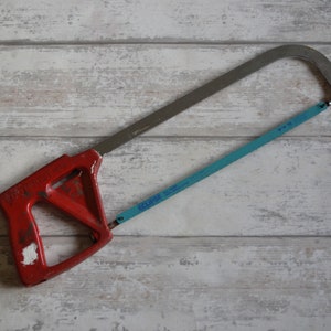 Puede incluir: Una sierra de mano roja y plateada con una hoja azul. La sierra tiene un mango de plástico rojo con forma triangular.