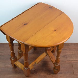 May include: A small, wooden, drop-leaf table with a half-circle top. The table is made of light brown wood and has four turned legs. The table is approximately 61 cm wide and 61 cm tall.