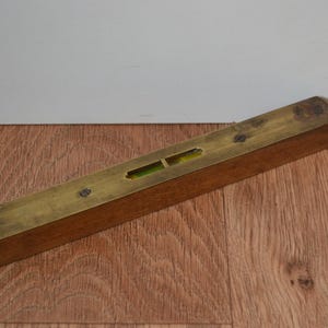 May include: A vintage wooden level with a brass top. The level has two small windows with a green liquid inside. The level is rectangular and has a few screws on the top. The level is resting on a wooden surface.