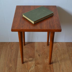 Könnte beinhalten: Ein quadratischer Beistelltisch aus Holz mit einer satten braunen Maserung und vier konisch zulaufenden Beinen. Ein rechteckiges Buch mit einem einfarbigen olivgrünen Einband liegt flach in der Mitte der Tischplatte und zeigt ein schlichtes Design.