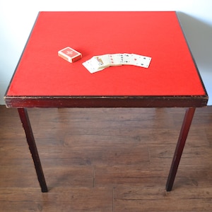 May include: A vintage card table with a bright red felt top and dark wooden legs. A deck of cards and a box of matches are on the table. The table is set on a wooden floor.
