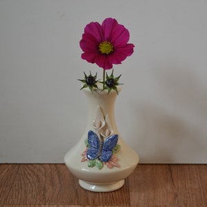 May include: A white ceramic vase with a butterfly design and a single pink flower.
