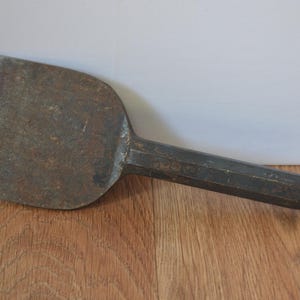 May include: A vintage metal chisel, showing signs of wear and tear, with a broad, rectangular blade and a long, hexagonal handle. The tool has a weathered, rusty appearance. The chisel is set against a wooden surface.