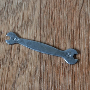 May include: A small, silver wrench with a curved handle and open-end heads. The wrench has the number "25" stamped on both ends. The tool is lying on a wooden surface.