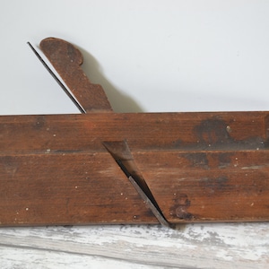 Può includere: Un pialla a mano in legno vintage con una lama in metallo. La pialla è marrone e ha una finitura usurata. È uno strumento tradizionale per la lavorazione del legno.