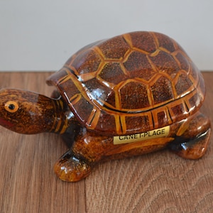Puede incluir: Figura decorativa de tortuga con acabado brillante. La tortuga tiene un caparazón marrón con un patrón geométrico, una cabeza y patas marrones. Una pequeña etiqueta dorada en el lateral dice "CANET-PLAGE".