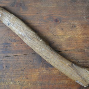 Peut inclure: Un manche en bois vieilli, probablement pour un marteau ou un outil similaire, est posé en diagonale sur une surface en bois sombre et texturée. Le manche est de couleur marron clair, avec des traces d'usure.