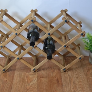 May include: A wooden wine rack with two bottles of wine. The rack is made of light-colored wood and has a crisscross design. The bottles are black with dark corks. A small potted plant is to the right.