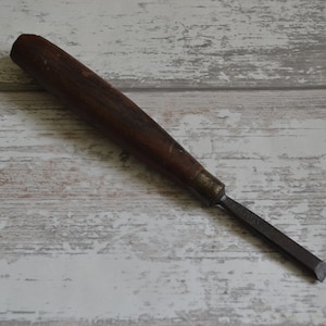 May include: A wooden-handled chisel with a metal blade. The chisel is lying on a white wooden surface.