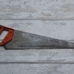 Puede incluir: Una sierra de mano con mango naranja y hoja de metal. La sierra está sobre una superficie de madera blanca.