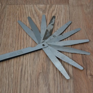 May include: A silver metal feeler gauge with a fan-shaped design. The gauge has multiple blades with different sizes marked on them. The gauge is labelled "GAY-1-5MM" and "BRITISH MADE".