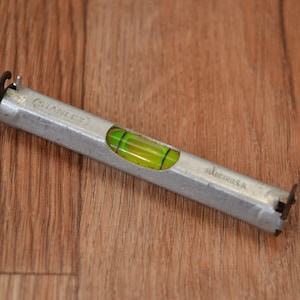 Può includere: Un livello Stanley argentato con un livello a bolla verde. Il livello è lungo circa 15 cm e ha un gancio a ciascuna estremità.