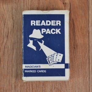 Puede incluir: Una baraja de cartas marcadas "Reader Pack". La caja de cartas es azul con texto y gráficos blancos, incluyendo una figura con sombrero y naipes. También está presente el texto "Magician's Marked Cards".