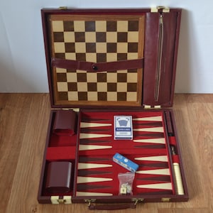 May include: Open burgundy leatherette game set with a chess board, backgammon board, playing cards, dice, and two brown leatherette cup holders. The set is open on a wooden surface.