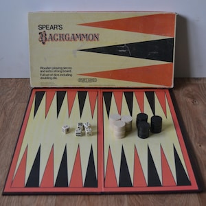 Puede incluir: Juego de Backgammon Spear's vintage. La caja es crema, naranja y negra. El tablero de juego abierto muestra un campo crema con triángulos negros y naranjas. Dados y fichas son visibles.
