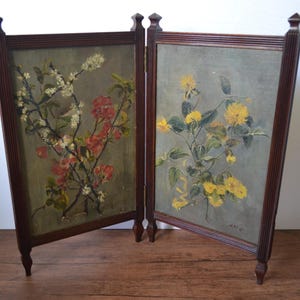 May include: A two-panel folding screen with dark wood frames and legs. Each panel features a floral painting, one with red and white blossoms, the other with yellow flowers. The background is a muted grey-green.