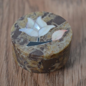 Puede incluir: Una caja redonda de mármol marrón y blanco con una decoración de incrustación de una flor blanca con una hoja rosa.