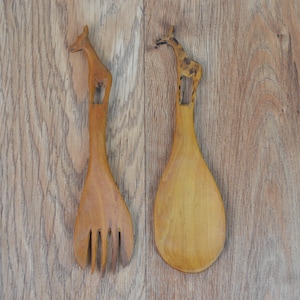 Puede incluir: Dos utensilios para ensalada de madera. Uno es un tenedor, el otro una cuchara. Ambos tienen mangos con forma de jirafa. El tenedor tiene cuatro púas. La cuchara tiene un cuenco redondeado. La madera es de color marrón claro.