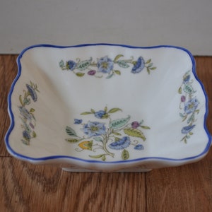 May include: A white square ceramic dish with a blue scalloped edge and a floral design in shades of blue, green, and yellow.