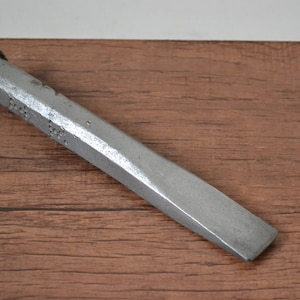 May include: A silver metal chisel with a textured head and a beveled edge. The chisel is lying on a wooden surface.