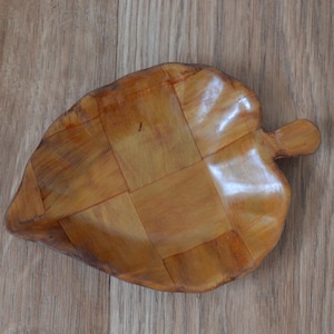 Op de afbeelding: Een houten kom in de vorm van een blad met een geweven patroon. De kom is gemaakt van lichtbruin hout en heeft een natuurlijke afwerking.