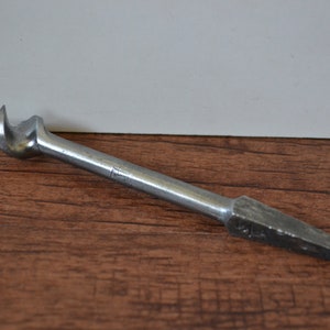 Puede incluir: Una broca de metal plateado con una punta de tornillo y un extremo plano y puntiagudo. La broca está sobre una superficie de madera.