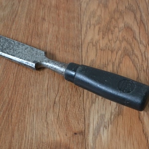May include: A silver metal chisel with a black plastic handle. The handle has a round, black logo on it.