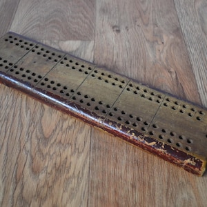 May include: A rectangular wooden cribbage board featuring a series of drilled holes arranged in rows for scoring. The board has a weathered, light brown surface with a dark, worn reddish-brown painted edge showing signs of age and use.