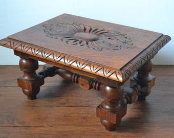 May include: A rectangular dark brown wooden footstool featuring a carved floral motif on the top surface and a decorative border. The stool is supported by four turned legs with square bases, connected by a central wooden stretcher frame.