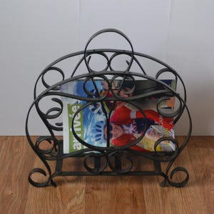 May include: A wrought iron magazine rack with a handle, holding two magazines. The rack has a decorative, scrollwork design and a dark finish. The magazines have colorful covers with images of people and text.