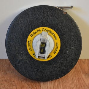 Puede incluir: Una cinta métrica negra y amarilla con el texto "Rabone Chesterman" y "Made in England" impreso en ella. La cinta métrica tiene un gancho de metal plateado y una funda de cuero negro.