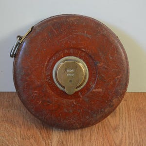 Puede incluir: Una cinta métrica de cuero marrón con una carcasa de latón. La cinta tiene 30,48 metros de largo y tiene el número "NF-501" impreso en ella.