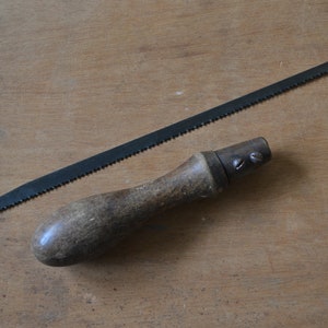 May include: A vintage saw with a wooden handle and a black metal blade. The saw is lying on a wooden surface.