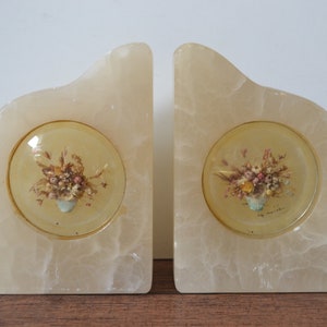 Puede incluir: Un par de sujetalibros de alabastro blanco con un inserto redondo de vidrio transparente que presenta un ramo de flores prensadas. Los sujetalibros tienen forma de triángulo curvo.