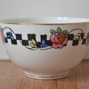 May include: White ceramic bowl with a black and white checkered pattern and floral design. The bowl has a gold rim and a gold band around the bottom.