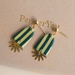 May include: A pair of gold earrings with green and yellow striped ceramic pendants. The pendants have a sunburst design at the bottom. The earrings are displayed on a box with the text "Pepper You" printed on it.
