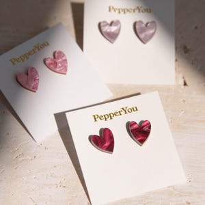 Könnte beinhalten: Drei Karten mit dem aufgedruckten Text "PepperYou". Jede Karte zeigt ein Paar herzförmiger Ohrringe. Die Ohrringe sind aus rosa und rotem marmoriertem Kunststoff.