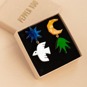 May include: A pair of mismatched earrings in a cardboard box labeled PEPPER YOU. One earring features a blue star and a white bird. The other earring features a yellow crescent moon and a green leaf-shaped charm, all connected by gold-tone metal.
