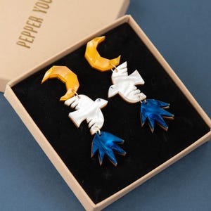 May include: A pair of dangle earrings featuring a yellow crescent moon, a white bird, and a blue tail section, presented in a tan box with the text PEPPER YOU printed on the lid. The earrings are set against a black velvet insert.