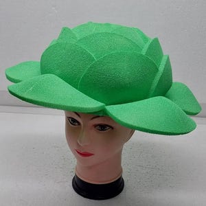 May include: A bright green, foam-like hat shaped like a large flower, worn by a mannequin head. The hat has multiple petals and a textured surface, creating a unique and eye-catching design. The mannequin head has a black base.