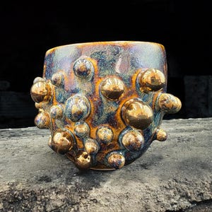 May include: A handmade ceramic mug with a distinctive textured finish. The mug is a mix of blue, brown, and gold hues, adorned with numerous gold-coloured, bubble-like features. The mug rests on a grey, textured surface.
