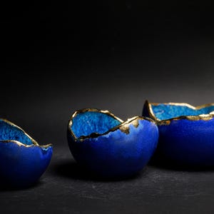 May include: Three blue ceramic bowls with gold-colored rims. The bowls are arranged in a row, each with a unique shape and size. The interior of the bowls has a lighter blue hue, creating a contrast with the exterior.