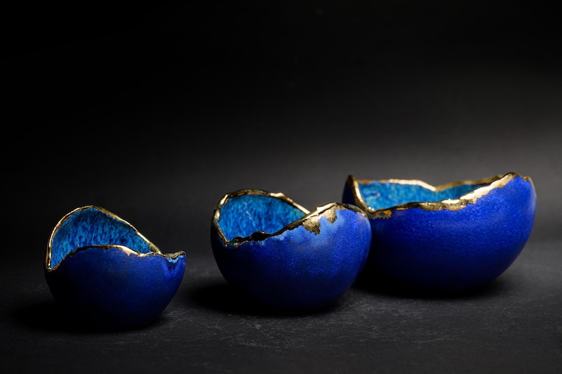 May include: Three blue ceramic bowls with gold-colored rims. The bowls are arranged in a row, each with a unique shape and size. The interior of the bowls has a lighter blue hue, creating a contrast with the exterior.