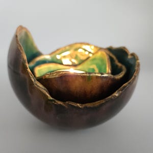 Puede incluir: Un juego de tres cuencos de cerámica anidados con una forma orgánica única. Los cuencos presentan un exterior marrón oscuro, un interior dorado y detalles en verde y turquesa. Los bordes tienen un diseño texturizado e irregular.