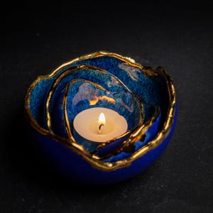 May include: A blue ceramic candle holder with a gold rim, holding a lit white tea light. The candle holder has a layered design, with the layers decreasing in size towards the center. The background is black.