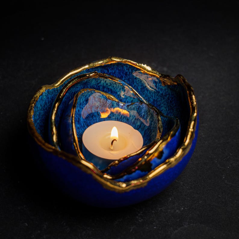 May include: A blue ceramic candle holder with a gold rim, holding a lit white tea light. The candle holder has a layered design, with the layers decreasing in size towards the center. The background is black.
