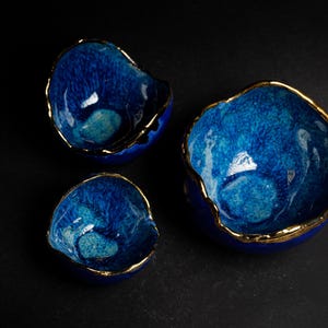 May include: Three blue ceramic bowls with gold rims. The bowls have a textured, crackled glaze and varying sizes. The bowls are arranged on a dark surface, showcasing their unique shapes and colors.