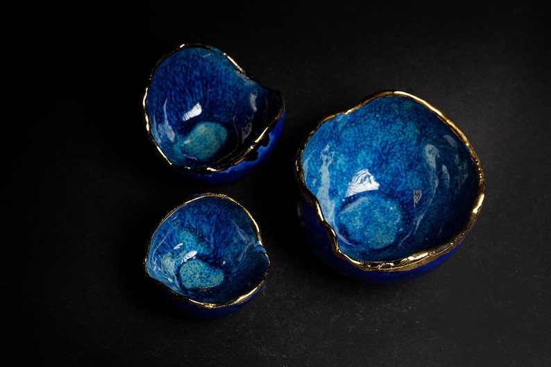May include: Three blue ceramic bowls with gold rims. The bowls have a textured, crackled glaze and varying sizes. The bowls are arranged on a dark surface, showcasing their unique shapes and colors.